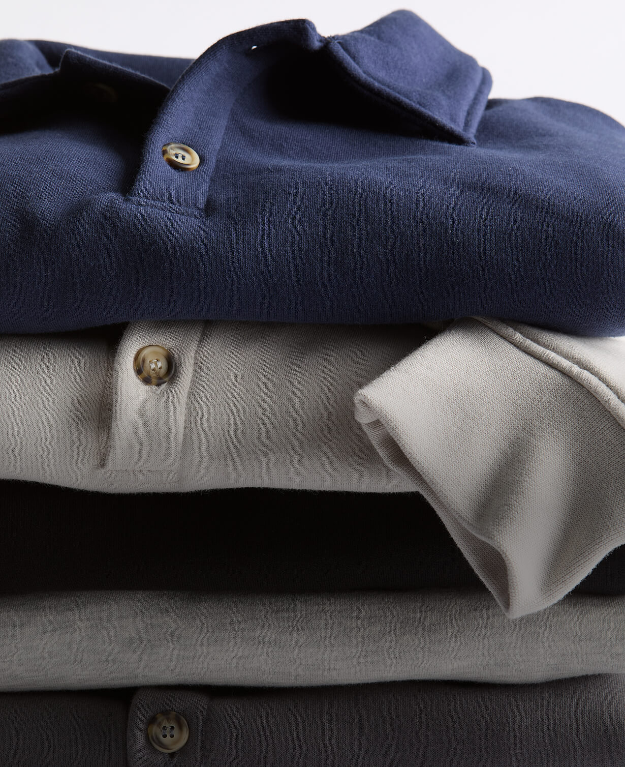 Stack of folded Unisex 10 oz Polo Sweatshirts in gray, black, cream, and navy, focused on the collar and horn buttons.