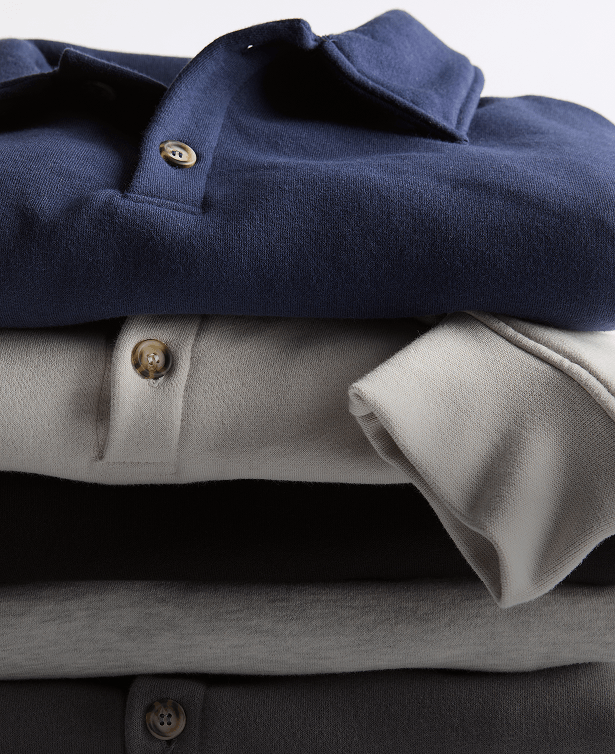 Stack of folded Unisex 10 oz Polo Sweatshirts in gray, black, cream, and navy, focused on the collar and horn buttons.