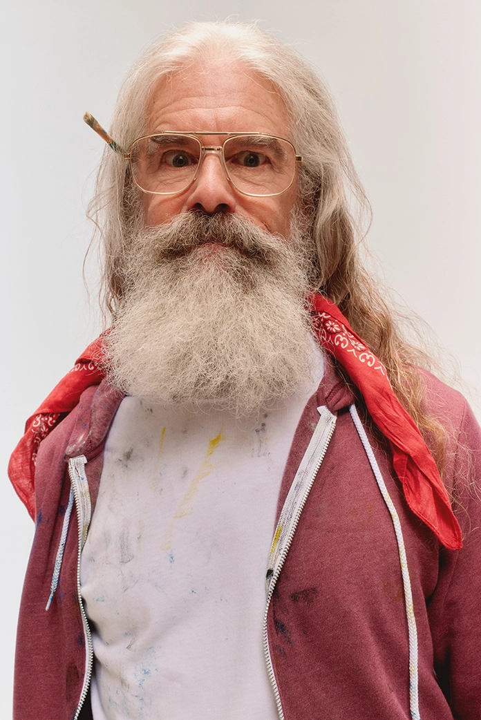 an older gentleman printmaker is looking directly at the camera, while wearing glasses, a paintbrush in his ear, and a zip-up BELLA+CANVAS hoodie in maroon
