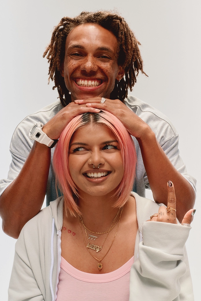 someone stands behind a seated girl with their hands on her head, both smiling